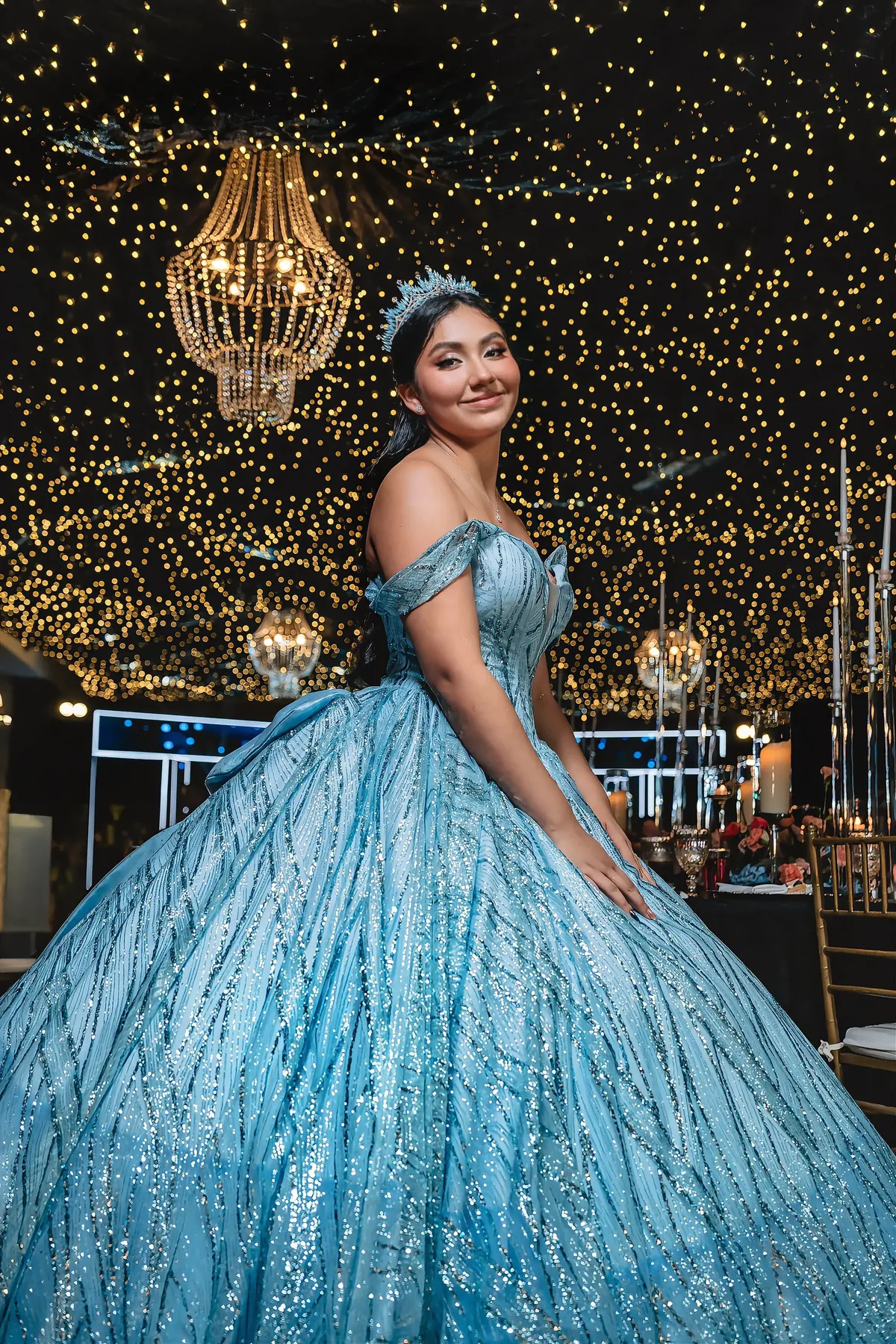 Quinceañera con vestido azul turquesa de cristales y tiara bajo candelabros con luces de hadas